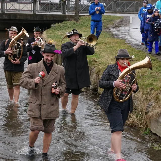 Musikanten mit Hut und hochgekrempelten Hosen spielen barfuß im Bach, am Ufer schauen Kinder zu