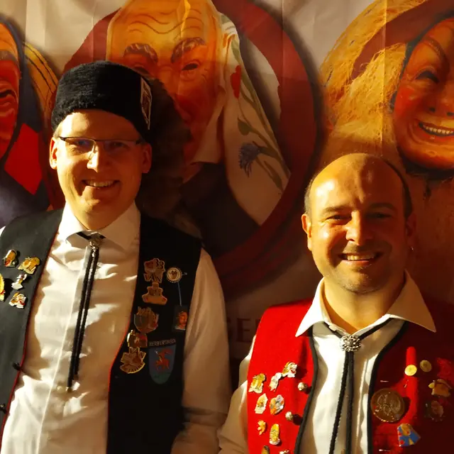 Bürgermeister Magnus Hoppe und Zunftmeister Stefan Schnitzer lachen vor einem Banner mit den Masken der Zunft