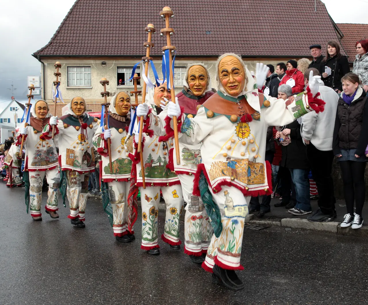 Eine Reihe Weckazwinger mit Schellenstäben beim Umzug, am Straßenrand viele Zuschauer