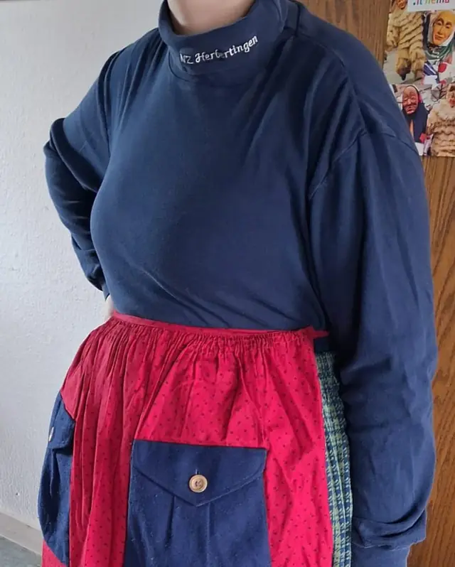 Blauer Rollkragenpullover mit Aufdruck der Narrenzunft, darüber die rote Schürze mit blauer Tasche