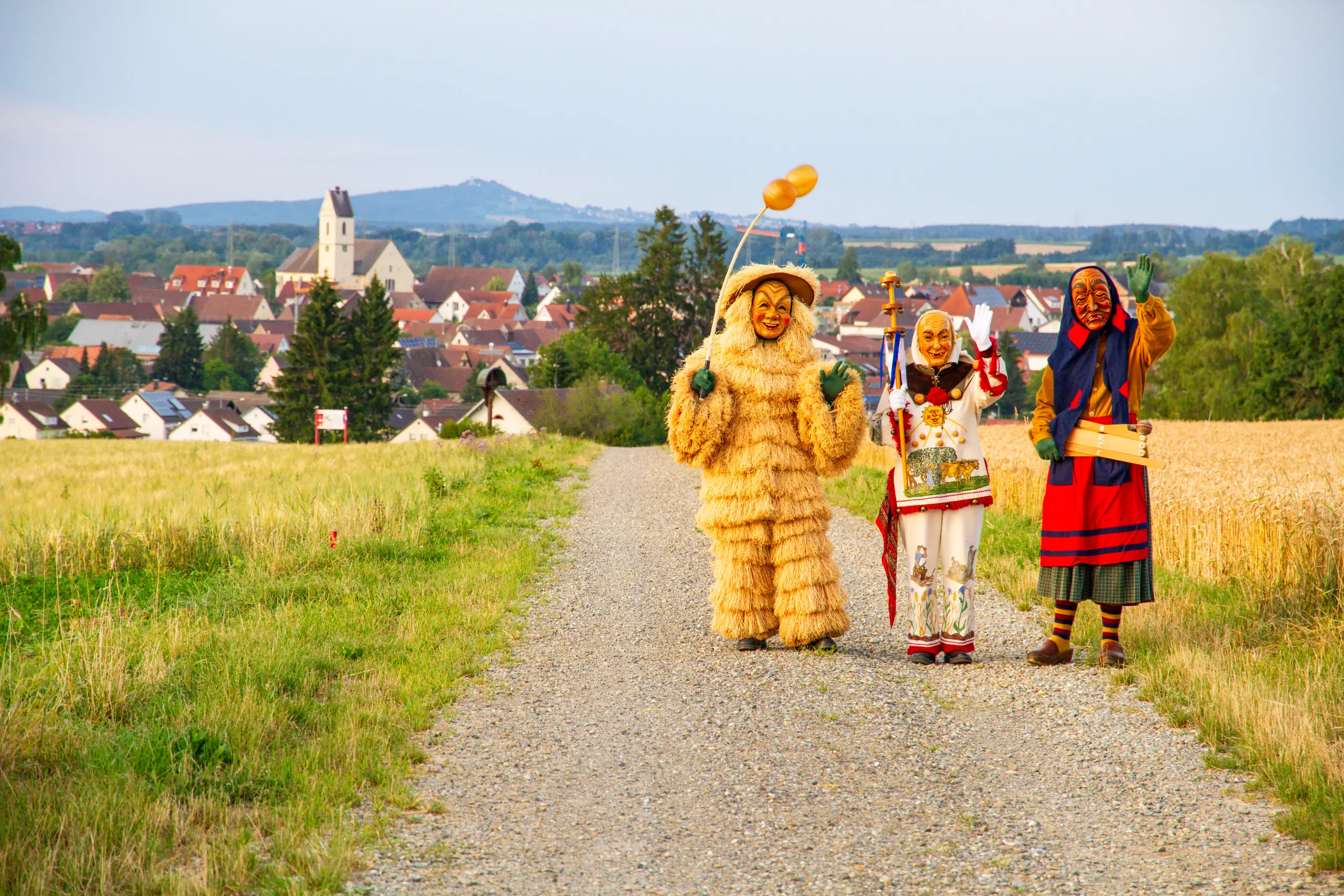 Strohbutz, Weckazwinger und Schrättele winken auf einem Feldweg, im Hintergrund Herbertingen mit Kirchturm